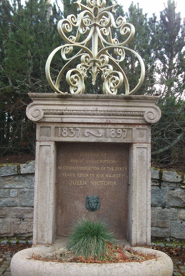 A stone memorial with a lion head below the text and a metal sculpture with swirls on top